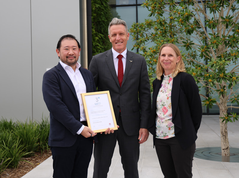 Andrew Thai stands holding a framed certificate with Josh Wilson in the middle and Magali Wardle to the right