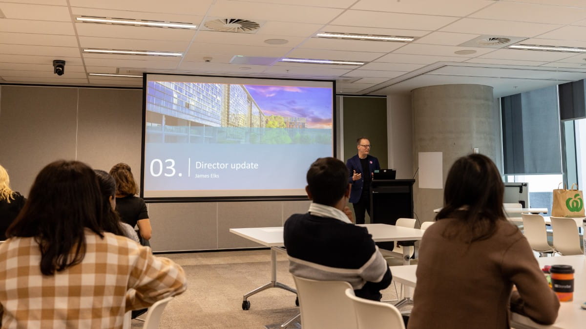 A mand stands at a podium talking beside a large screen with people sitting in front of him listening