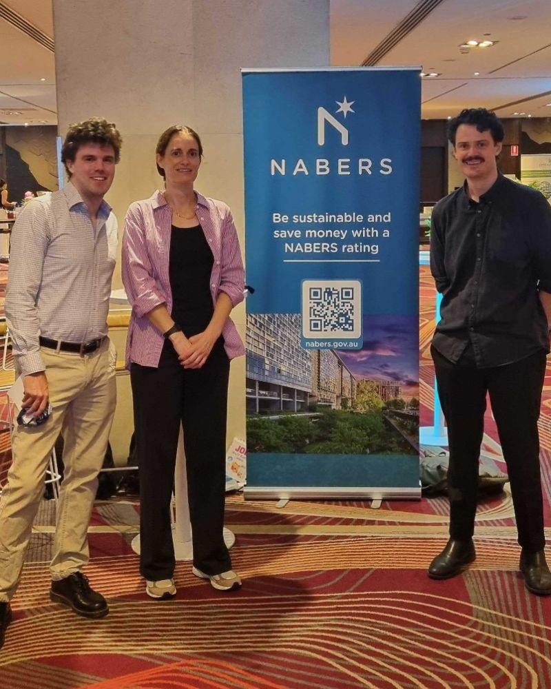 Three members of the NABERS team stand smiling at the conference centre with a NABERS pullup banner in the background
