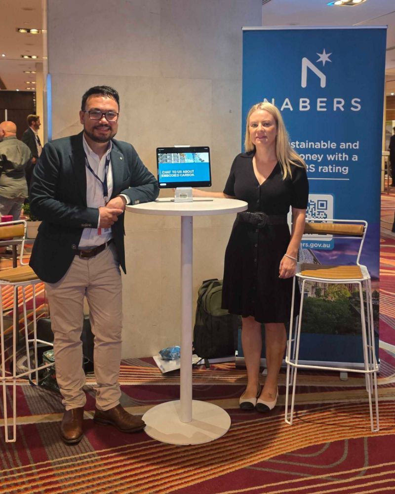 Two members of the NABERS team stand smiling beside a tall table at the conference centre